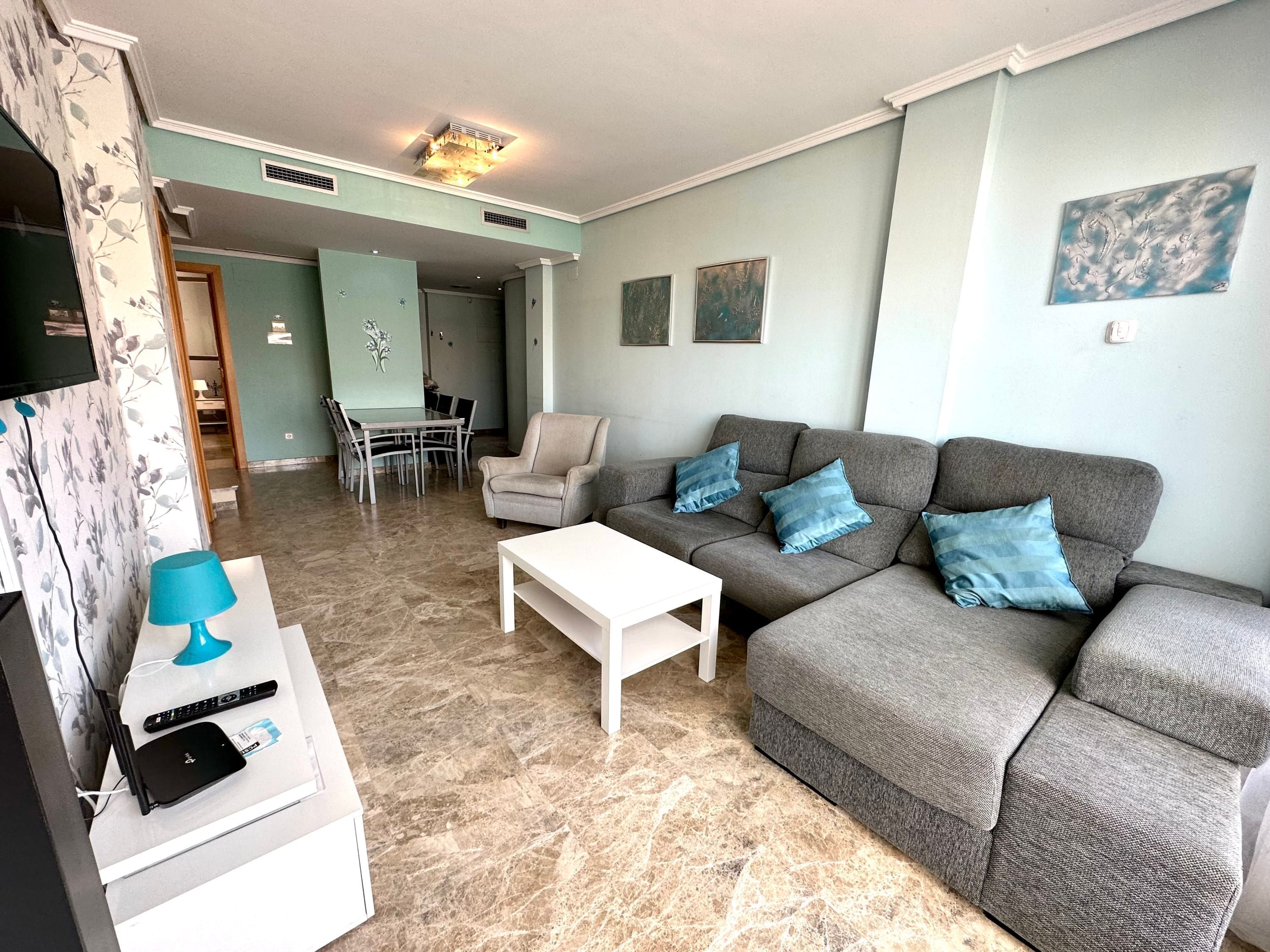 Un salón con un sofá gris, una mesa de centro blanca, una lámpara azul y un televisor sobre un mueble.