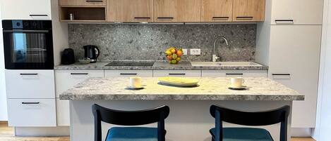 A modern kitchen with white cabinets, a granite countertop, a central island with two chairs, a hanging pendant light, and a built-in oven.