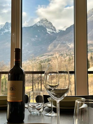 A bottle of wine and two glasses on a table with a view of mountains through large windows.