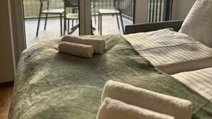 Une chambre avec une couvre-lit verte, deux serviettes enroulées et une vue sur un balcon agrémenté de chaises et d’une table.