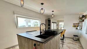 A modern kitchen with a central island, black countertop, and a sink. There are hanging pendant lights, a window with a view, and a tiled floor. - Casa Con Vista - Stunning Villa in Moraira (Teulada)