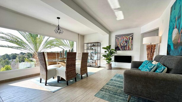 A modern living room with a glass table, wicker chairs, a grey sofa, and a large window offering a view of palm trees and hills. - Casa Con Vista - Stunning Villa in Moraira (Teulada)