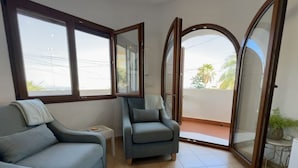 Un salon comprenant un canapé, un fauteuil et une petite table. De grandes fenêtres et une porte-fenêtre coulissante donnent sur un balcon.