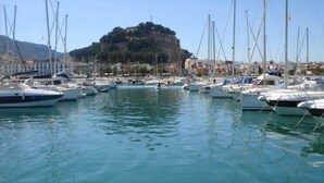 Un port de plaisance où sont amarrés de nombreux bateaux, une colline imposante avec des bâtiments en arrière-plan et une eau calme.