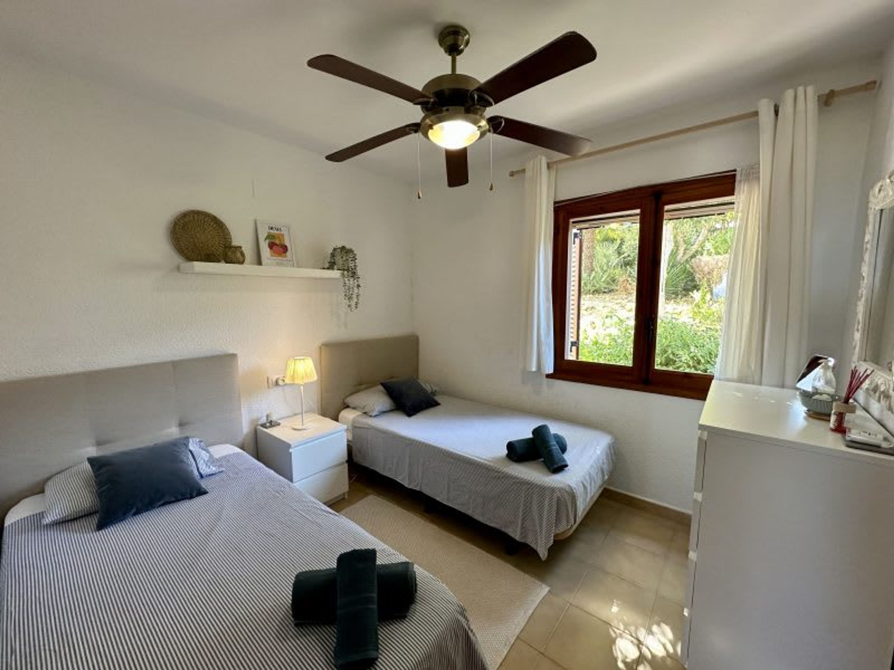 Una habitación con dos camas, un ventilador de techo, una ventana con cortinas y una pequeña mesa con una lámpara.