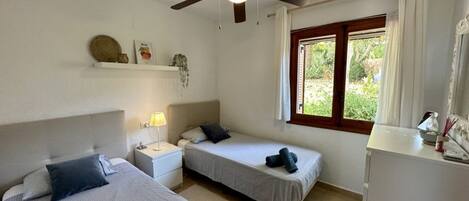 Una habitación con dos camas, un ventilador de techo, una ventana con cortinas y una pequeña mesa con una lámpara.