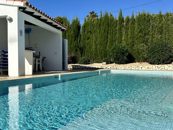 Una piscina con zona de solárium con baldosas, área de descanso techada y pared de piedra con árboles al fondo.
