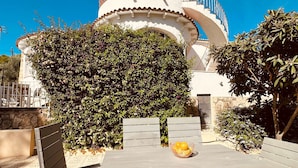 Un área de estar al aire libre con una mesa y sillas, rodeada de vegetación y un edificio con fachada blanca y detalles decorativos rojos.