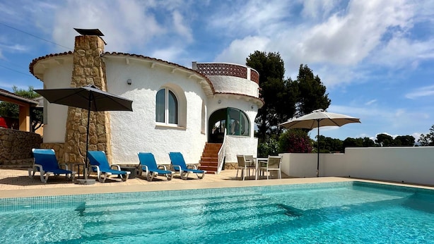 Una villa blanca con piscina, sillas de descanso azules y una chimenea de piedra.