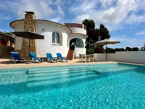 Una villa blanca con piscina, sillas de descanso azules y una chimenea de piedra.