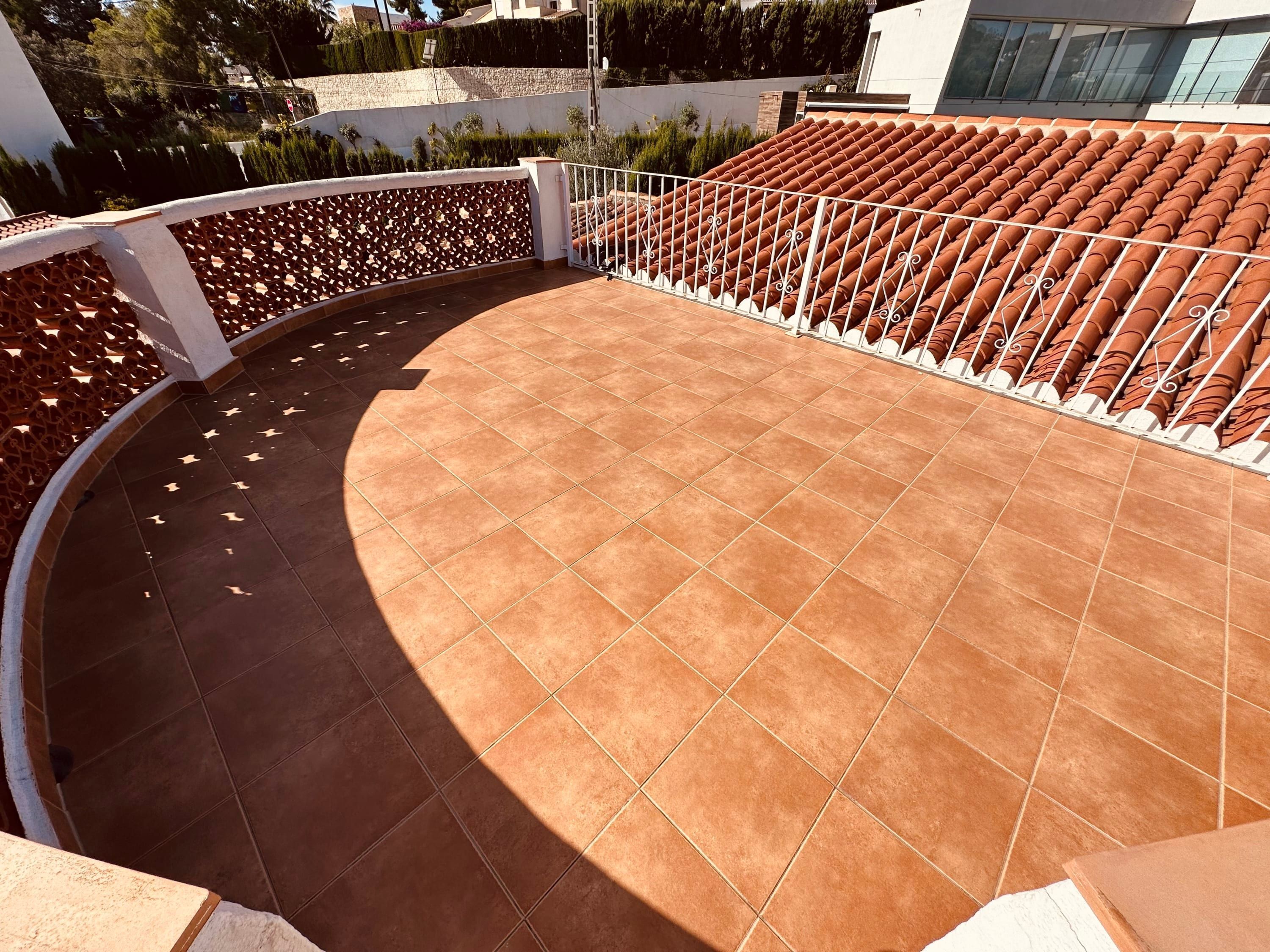 En buet udendørs terrasse med terracottafliser, hvide gelændere og udsigt til et boligområde.