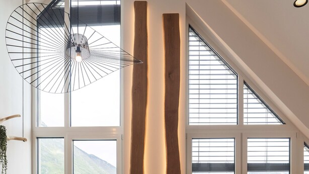 A modern living room with a wooden floor, a leather rug, a sofa, and a table. Large windows offer a view of mountains and a town. - So a Valley Suites - Superior Apartment 28 (Bovec)