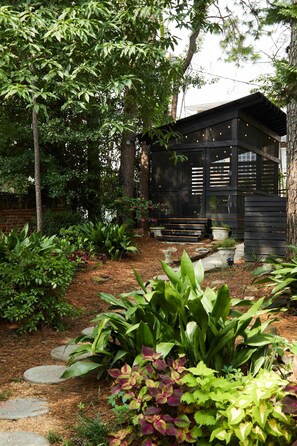 Une cabane en bois dotée d’un porche couvert, entourée d’un jardin où poussent différentes espèces de plantes et bordée d’un sentier en pierre.