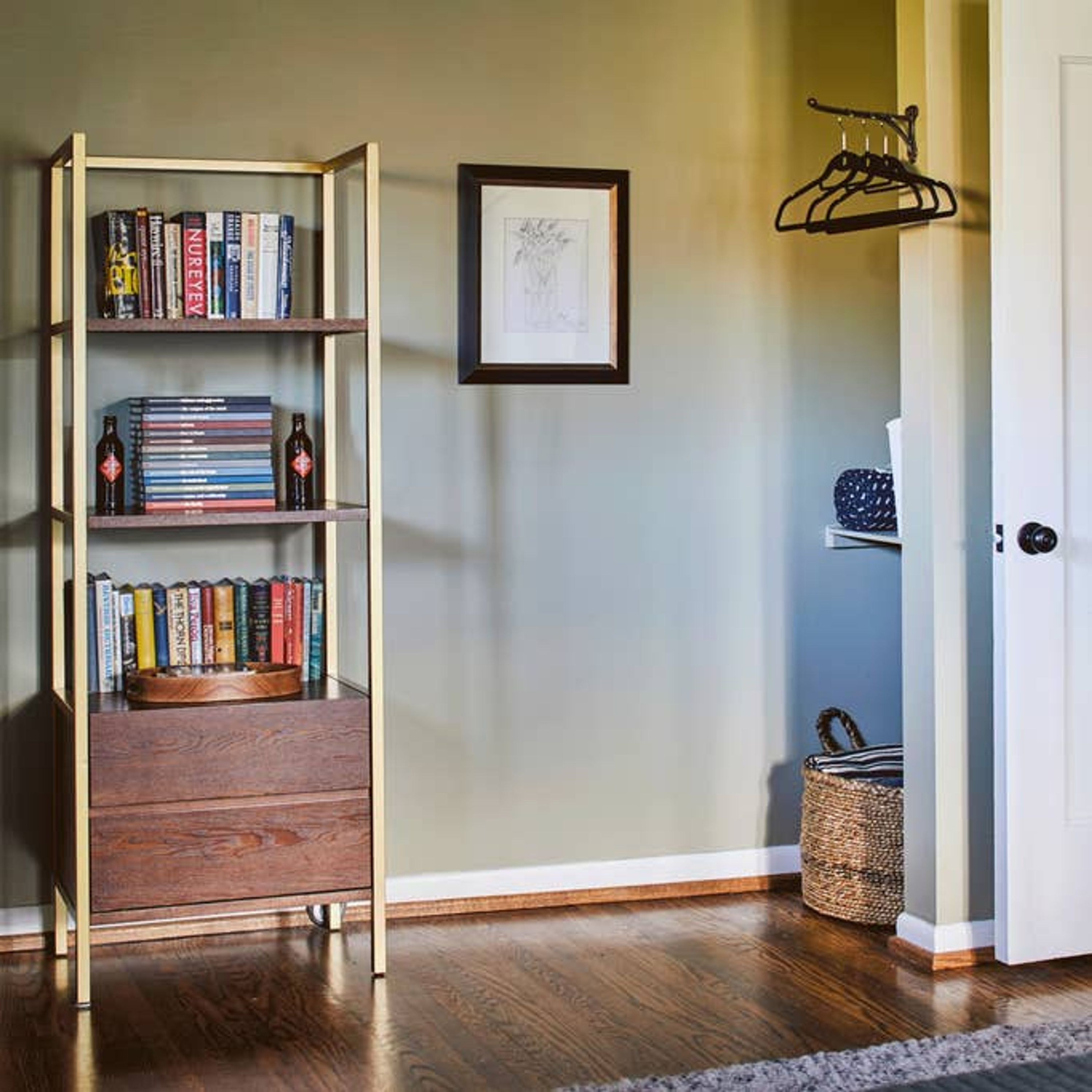 A room with a wooden floor, a bookshelf, a framed picture on the wall, a coat rack, and a basket.