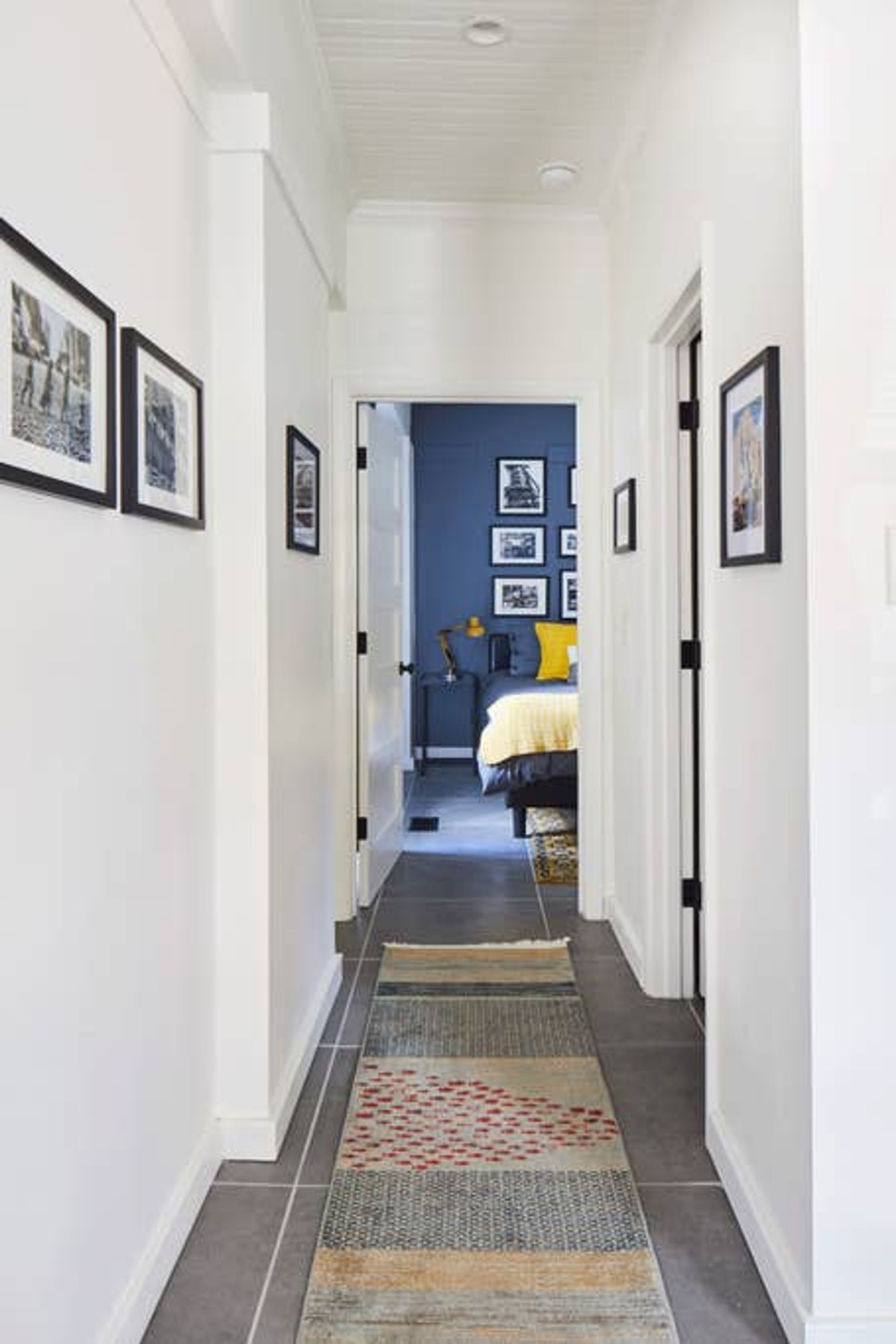 A hallway with framed pictures on the walls, leading to a bedroom with a bed and a yellow pillow.