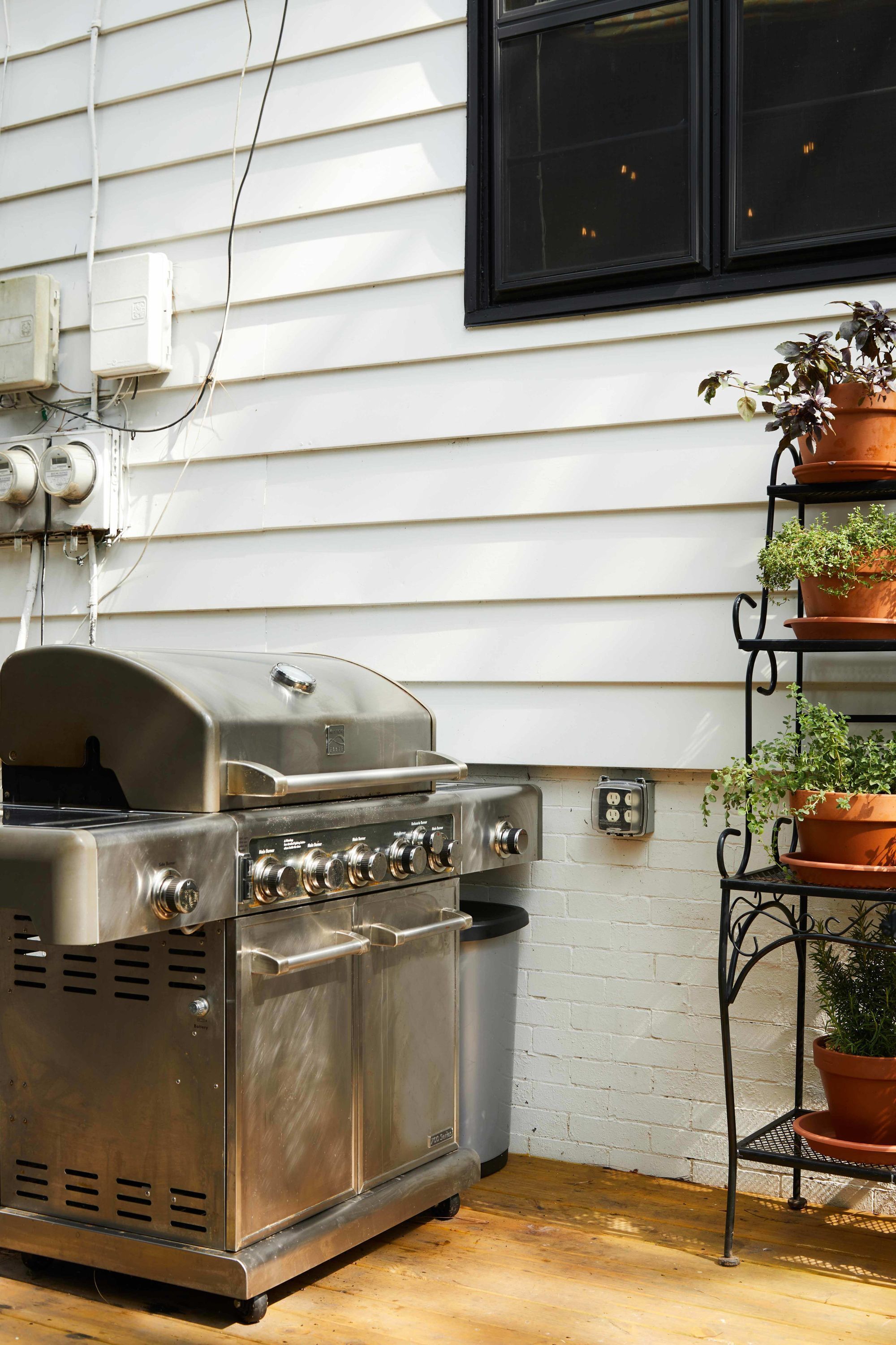 Una parrilla de acero inoxidable para exteriores con chimenea, un cubo de basura negro y un soporte de metal para plantas con macetas, todo frente a una pared blanca con una ventana arriba.