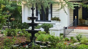 A white building with a stone fountain in the garden, surrounded by greenery and a wooden deck.
