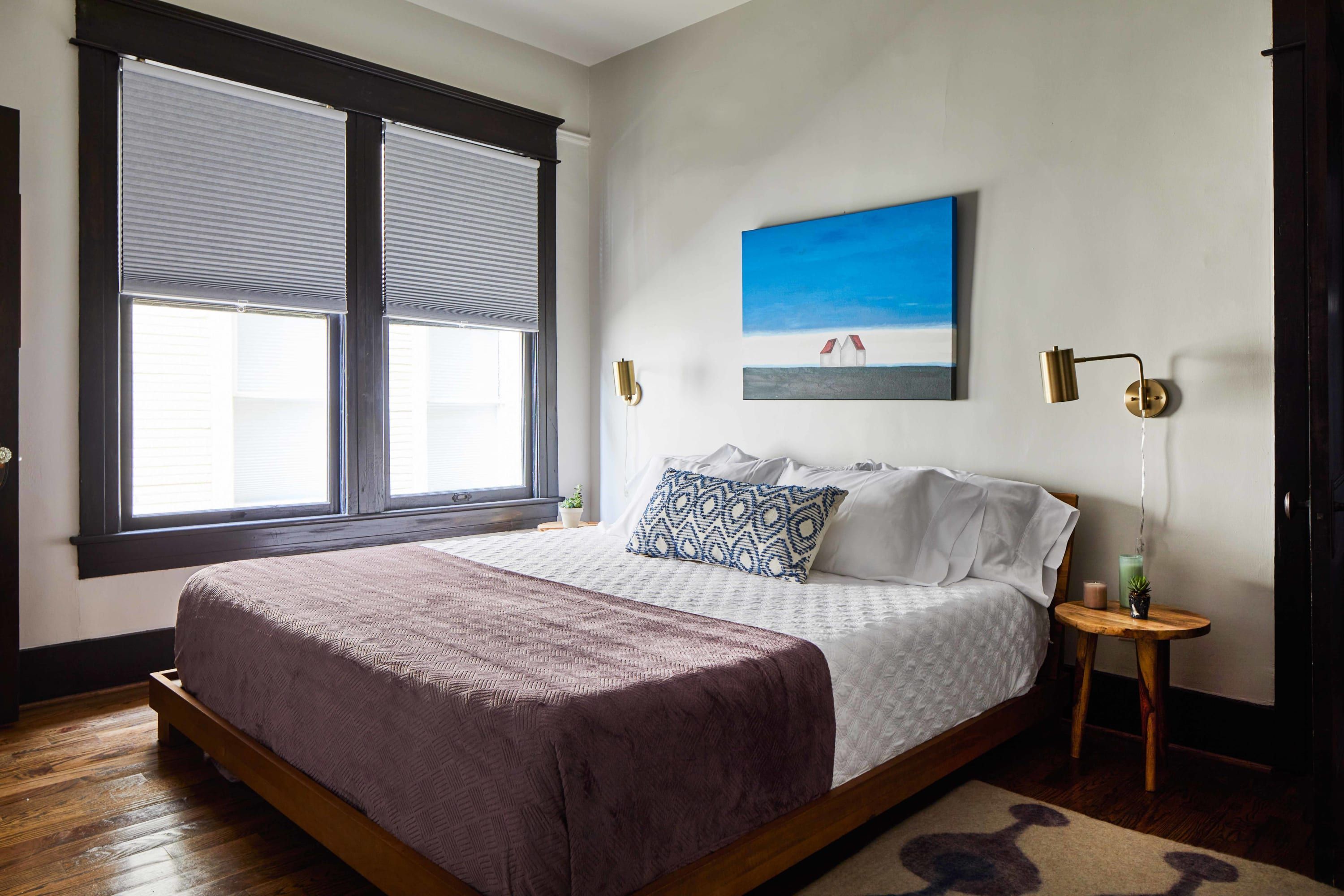 Un dormitorio con una cama grande, una ventana con persianas, una lámpara fijada a la pared y un cuadro.