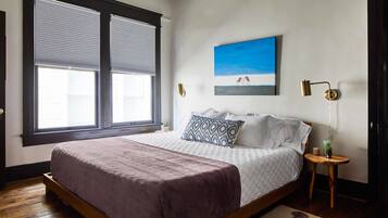 Un dormitorio con una cama grande, una ventana con persianas, una lámpara fijada a la pared y un cuadro.