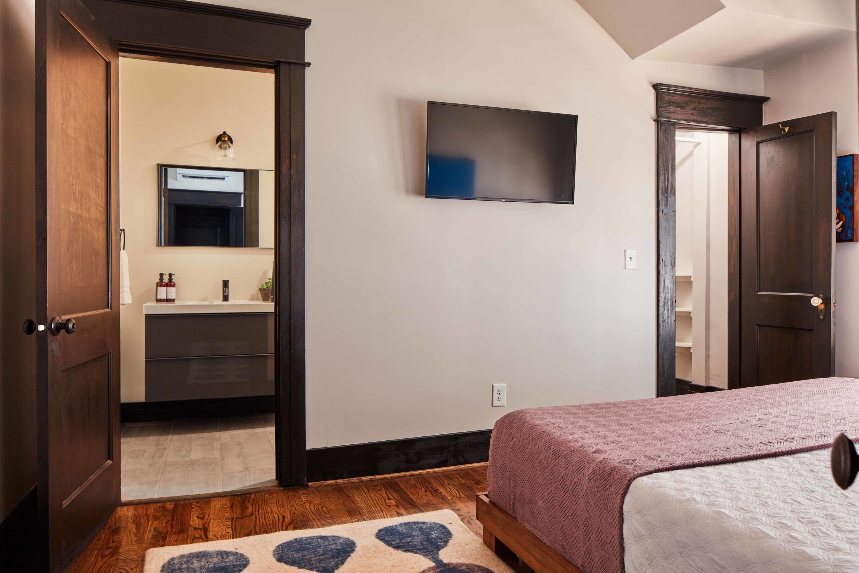 A bedroom with a bed, a flat-screen TV mounted on the wall, a wooden door, and a bathroom visible through an open door.