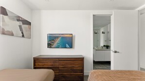 A bedroom with a wooden dresser, a painting of a beach, and a view into the bathroom.