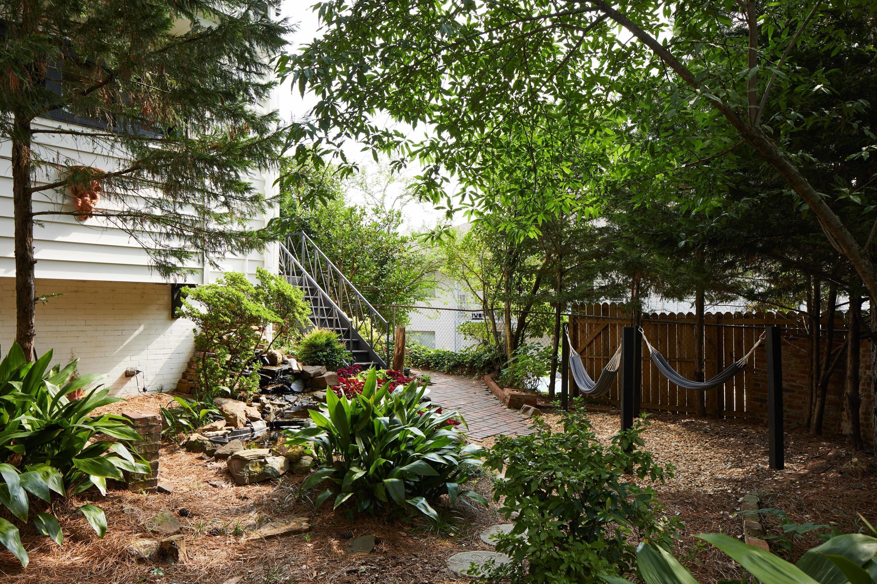 Un jardín con una escalera de madera que lleva a una casa, rodeado de diversas plantas y una hamaca.
