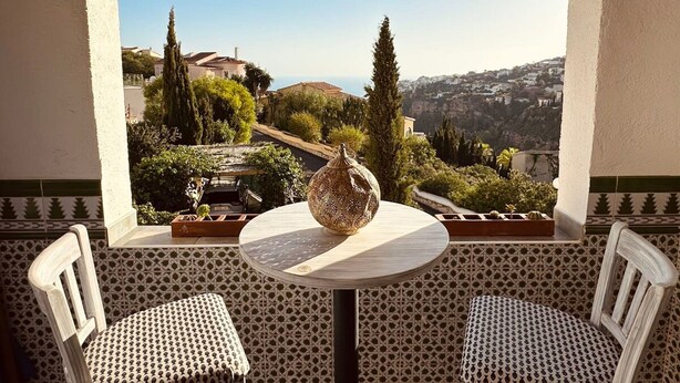 Un balcón con una mesa y dos sillas, con vista a un paisaje escénico de una colina con árboles y edificios.