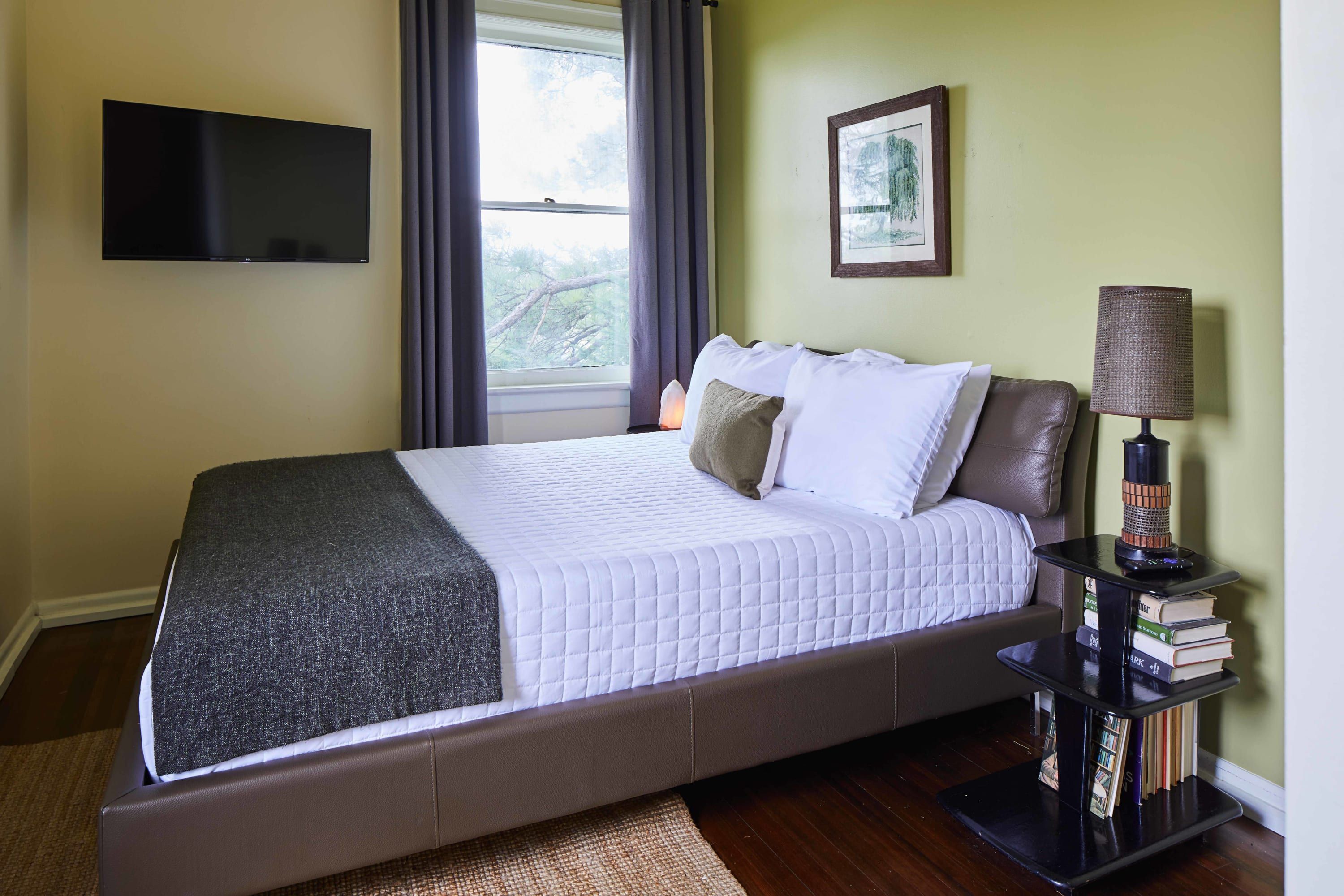 A neatly made bed with a white quilt, a dark blanket, and several pillows. A flat-screen TV is mounted on the wall above the bed. There is a window with curtains, a framed picture on the wall, and a small table with a lamp and books beside the bed.