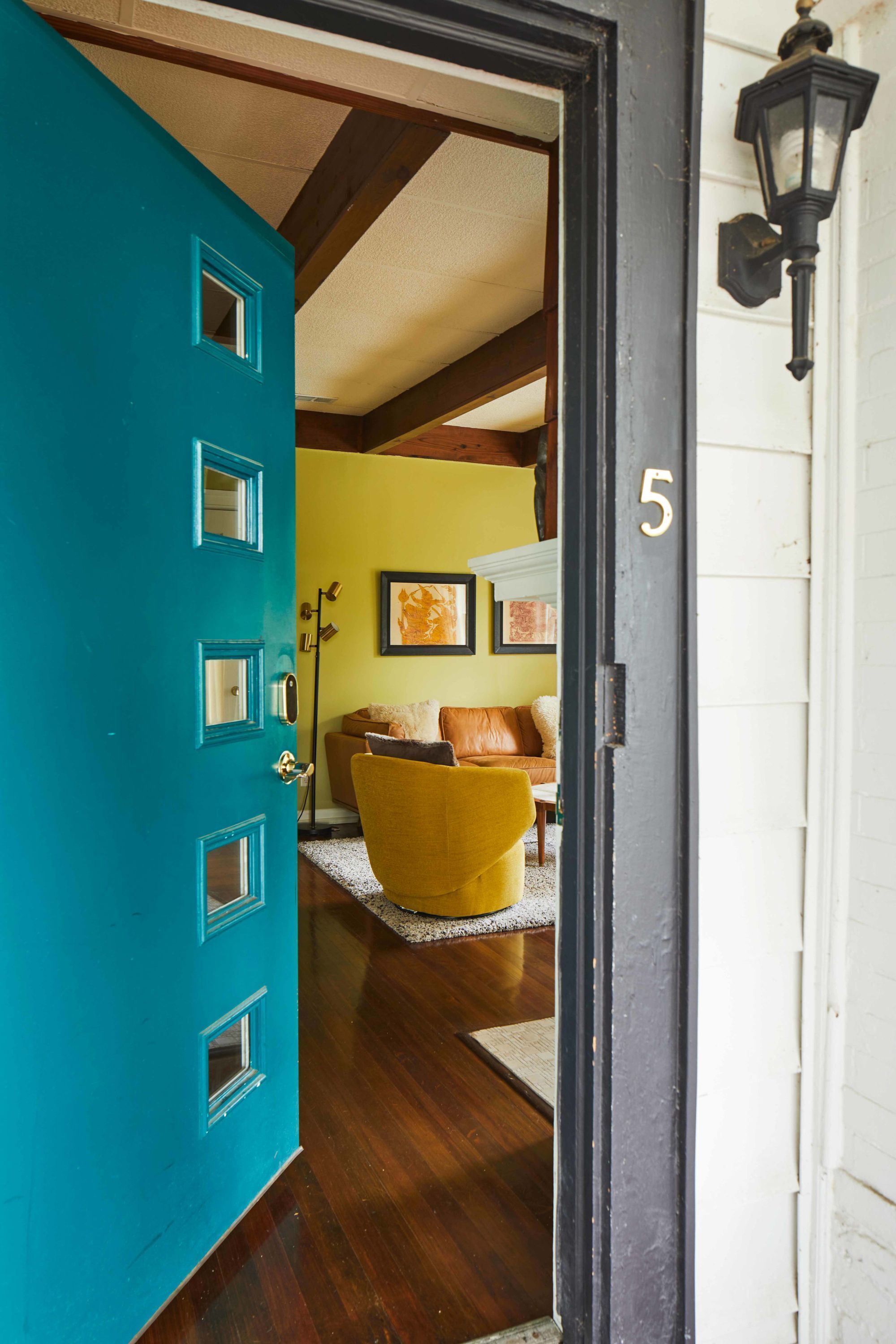 Uma porta azul-turquesa com o número '5' na parede, que dá acesso a um quarto com um sofá amarelo e obras de arte emolduradas.