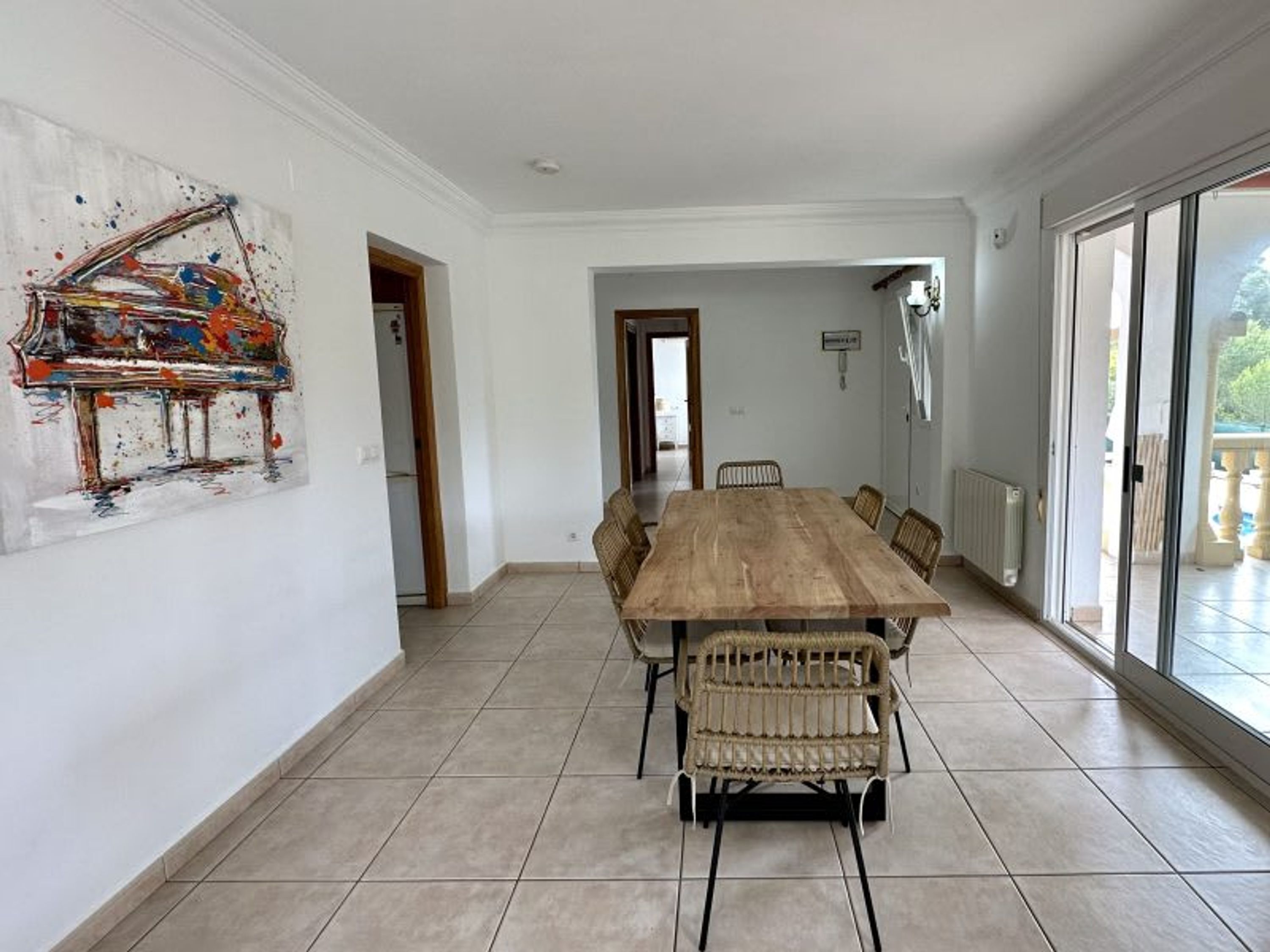 Un comedor amplio con una gran mesa de madera y sillas de mimbre, una pared blanca con un cuadro abstracto colorido y vistas a través de un ventanal.