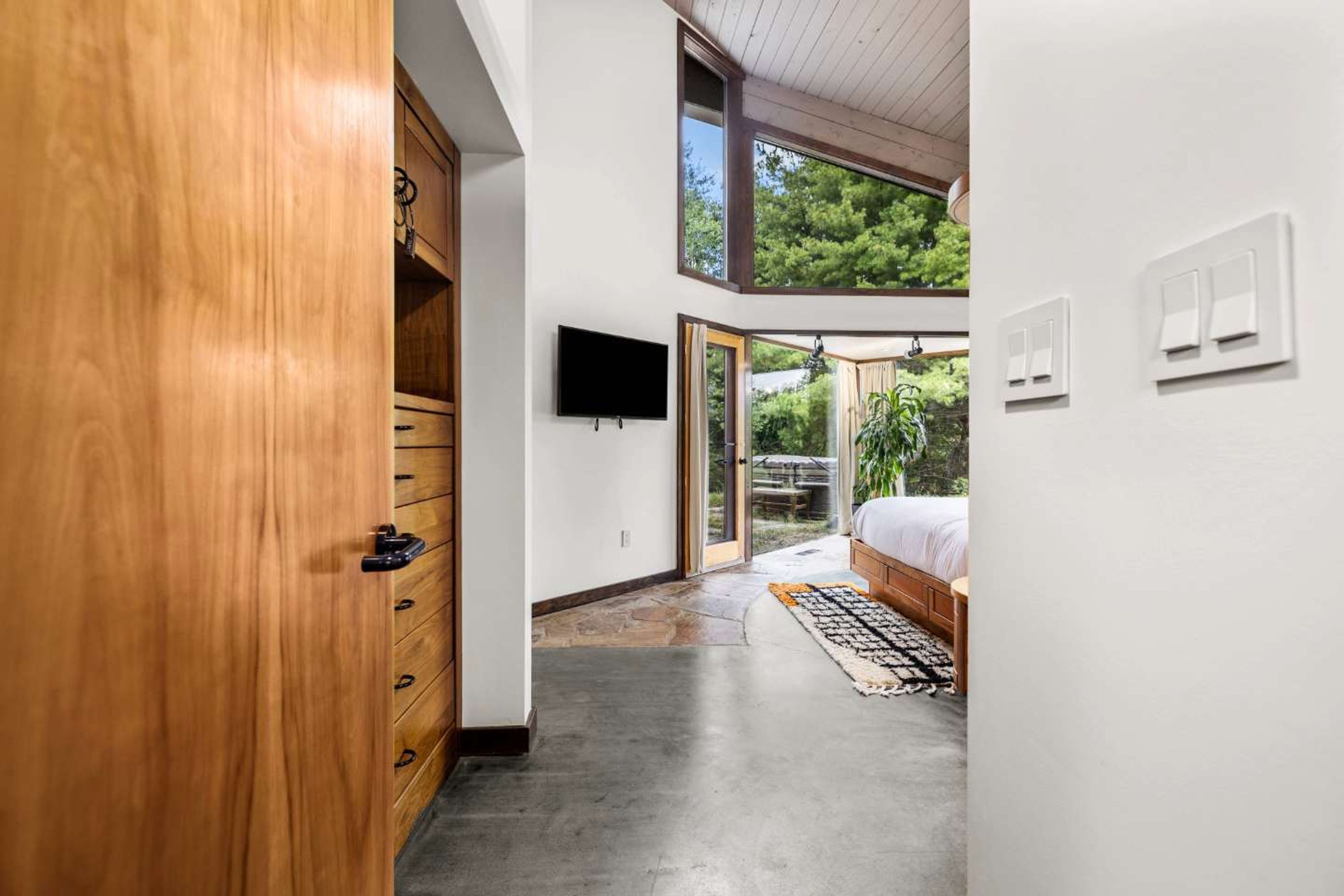 A modern bedroom with a large bed, a TV mounted on the wall, and a view of the outdoors through a sliding door.