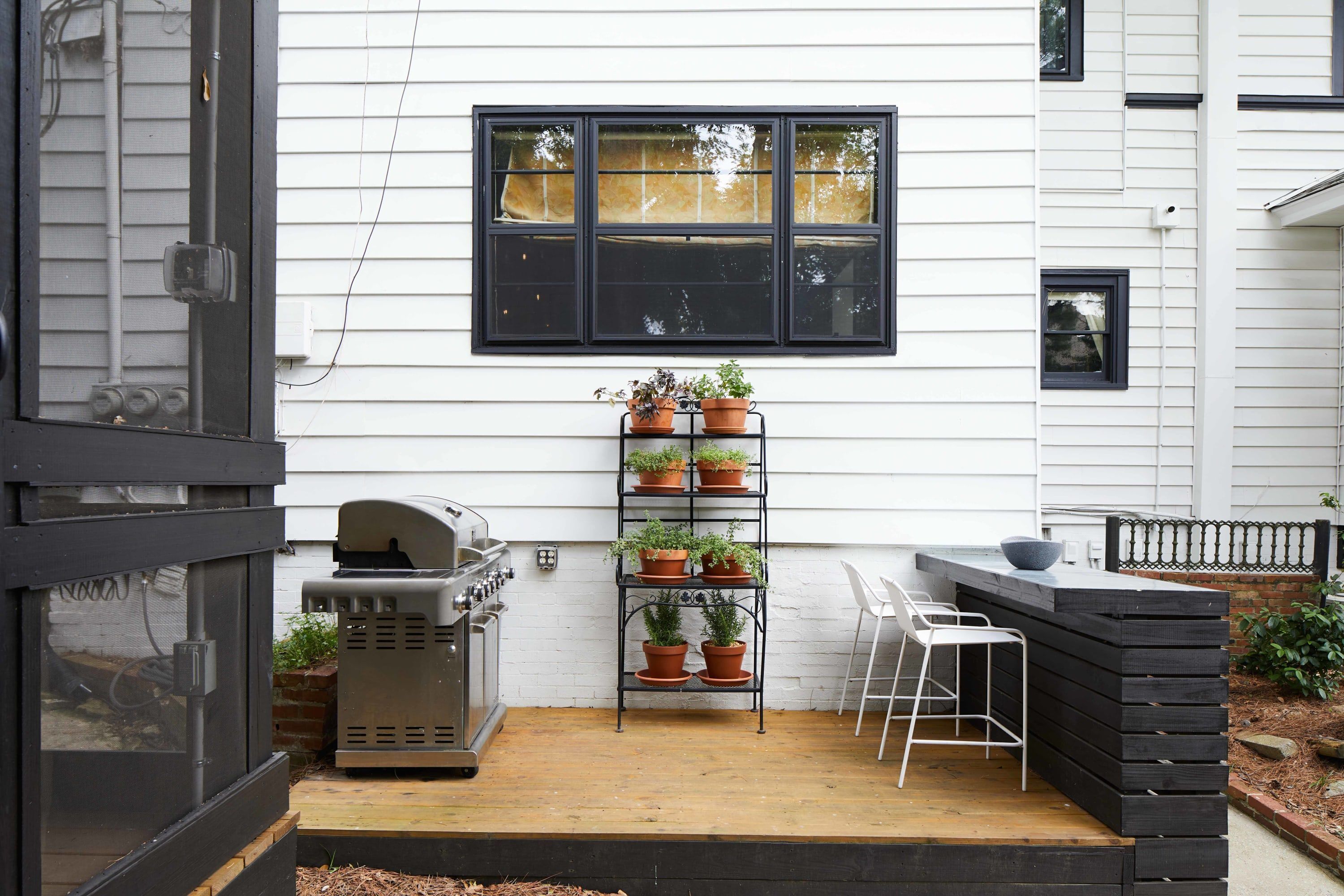 A patio with a grill, a potted plant shelf, and a chair.