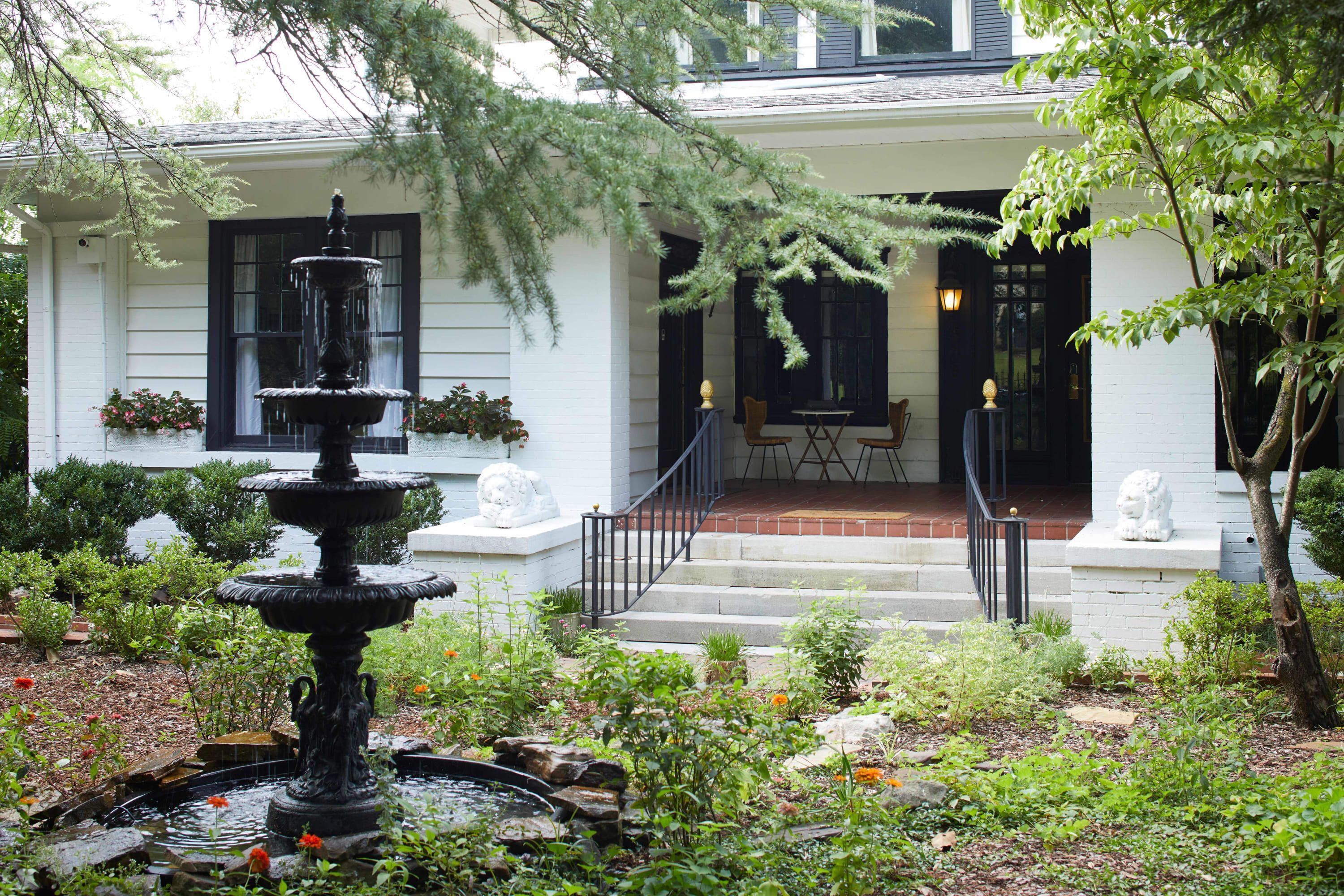 A white building with a fountain, steps leading to a porch, and a garden with various plants and flowers.