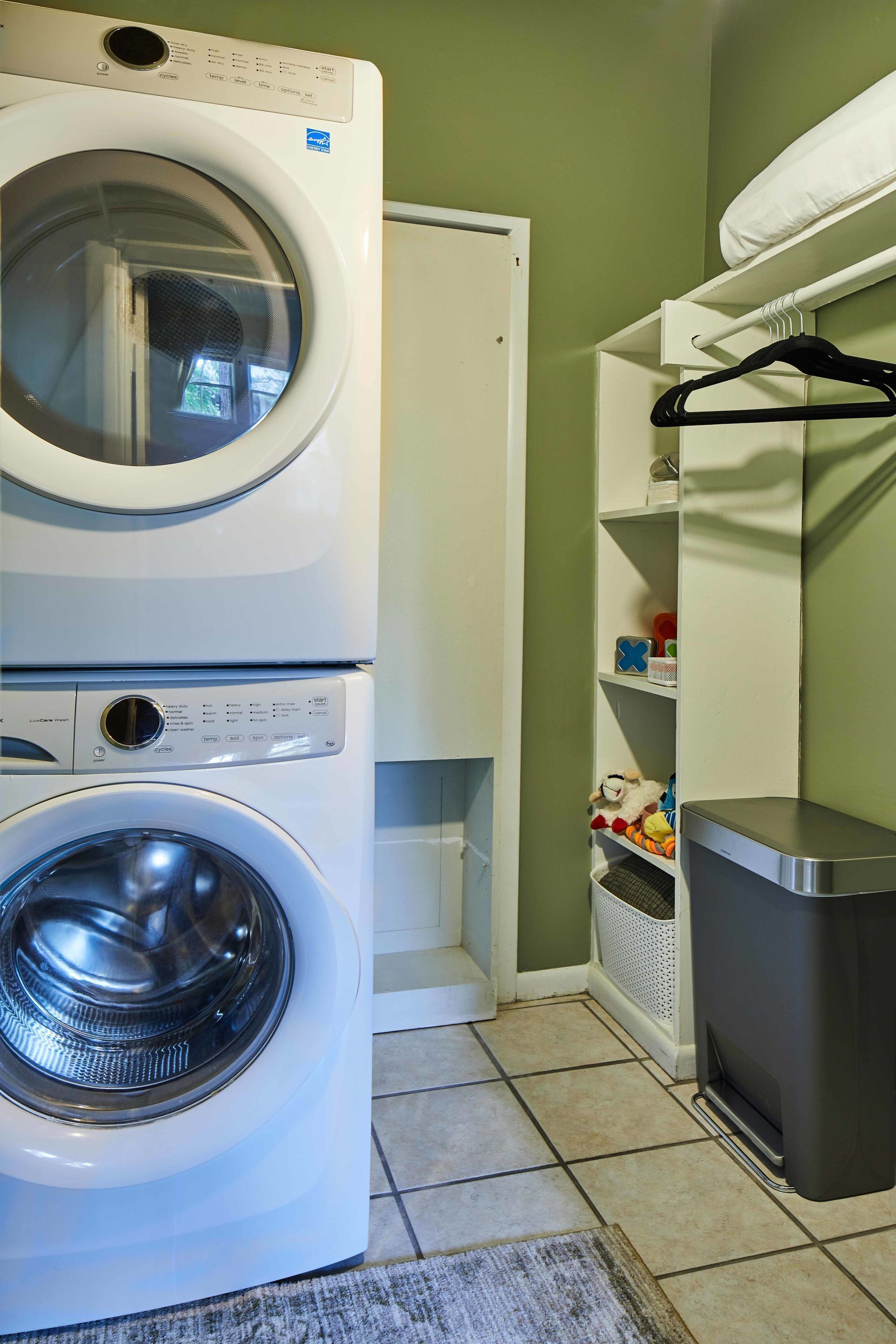 A laundry area with two stacked washing machines, a shelf with toys, and a trash bin.