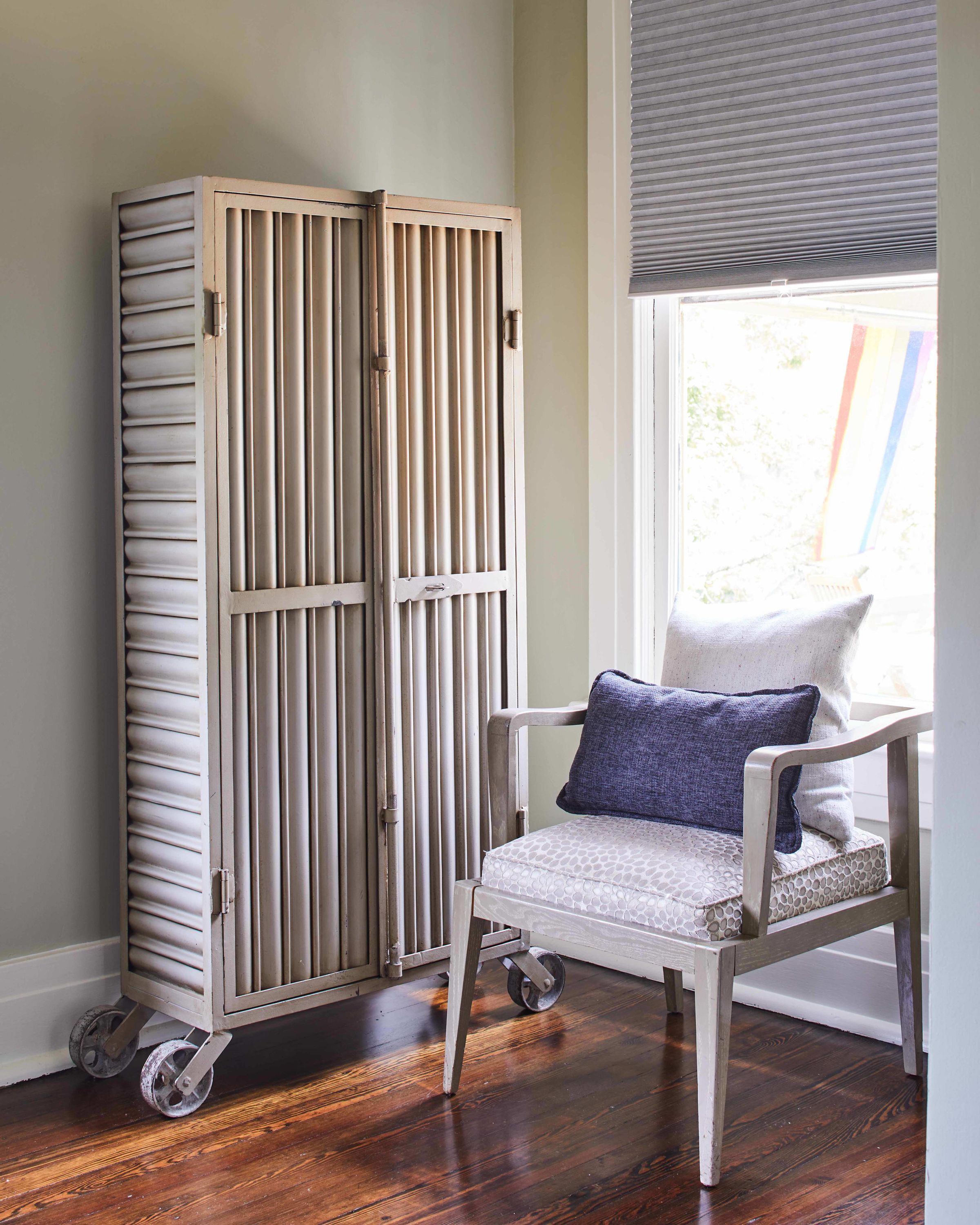 A room with a wooden wardrobe, a chair with a cushion, and a window with blinds.