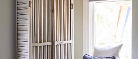 A room with a wooden wardrobe, a chair with a cushion, and a window with blinds.