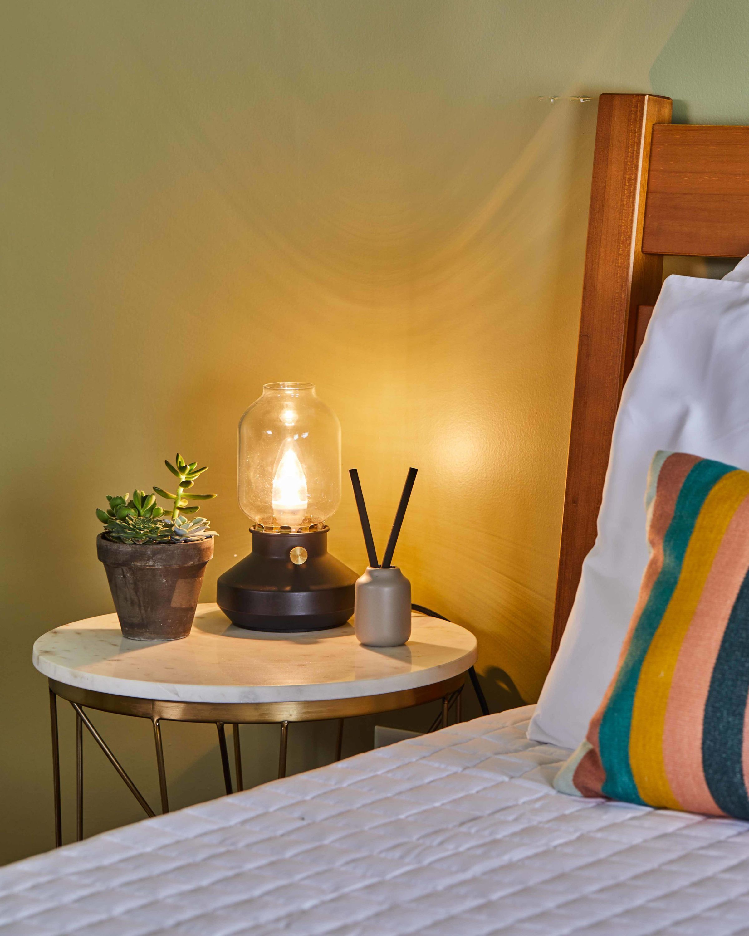 A neatly made bed with a striped pillow, a small table with a lamp, a potted plant, and a vase with reed diffusers.