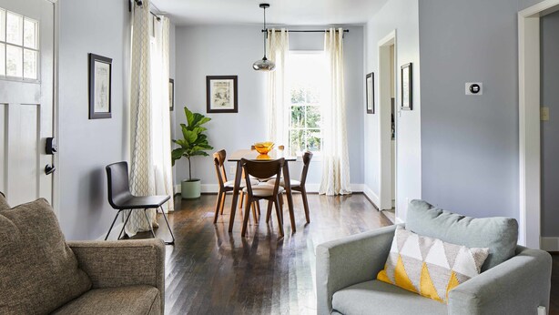 Un salon avec un canapé gris, un fauteuil marron et une table basse en bois. On y trouve également un coin repas avec une table et des chaises, une plante en pot et une fenêtre avec des rideaux.