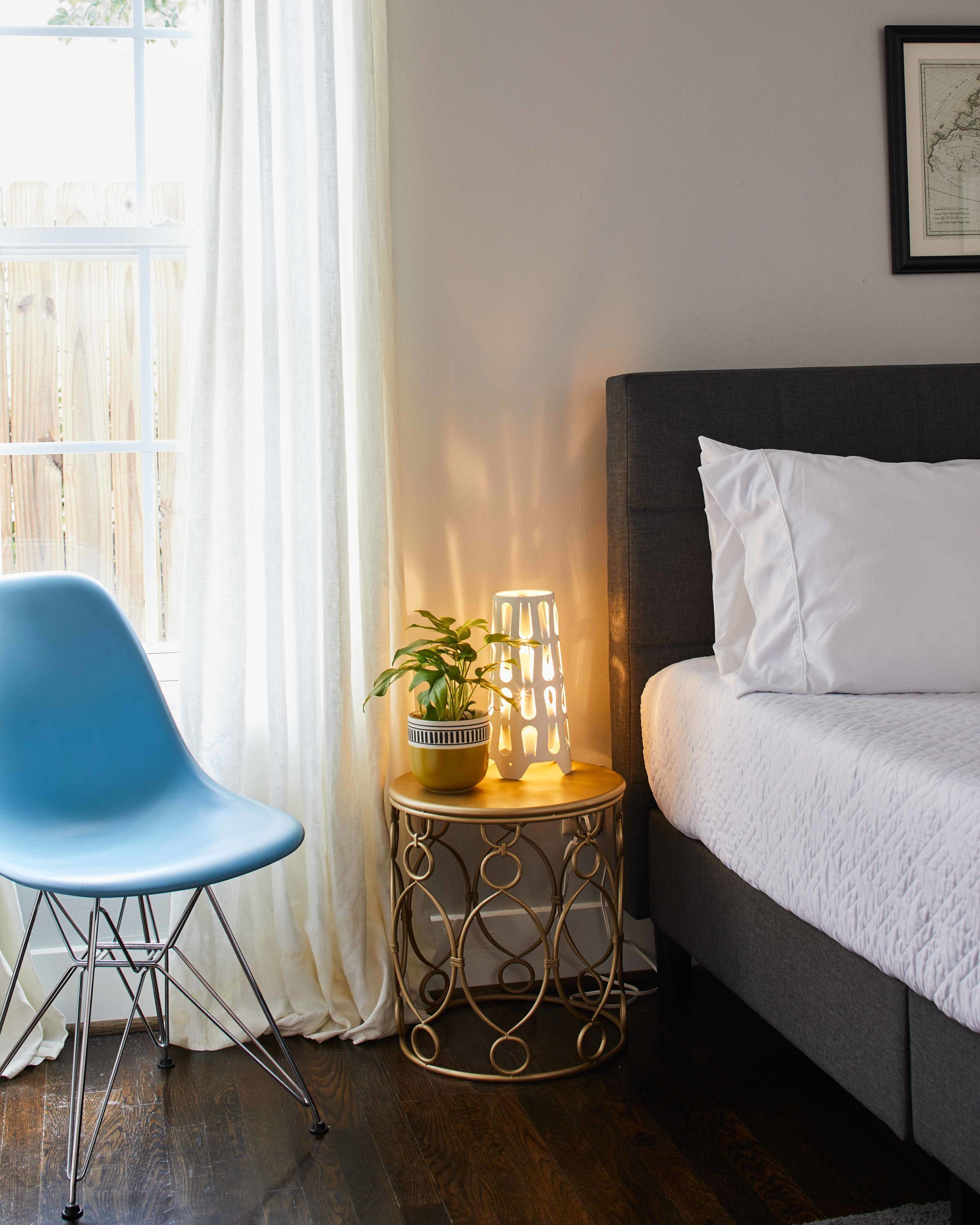 Een slaapkamer met een bed, een blauwe stoel, een kleine tafel met een plant en een lamp, en een raam met gordijnen.