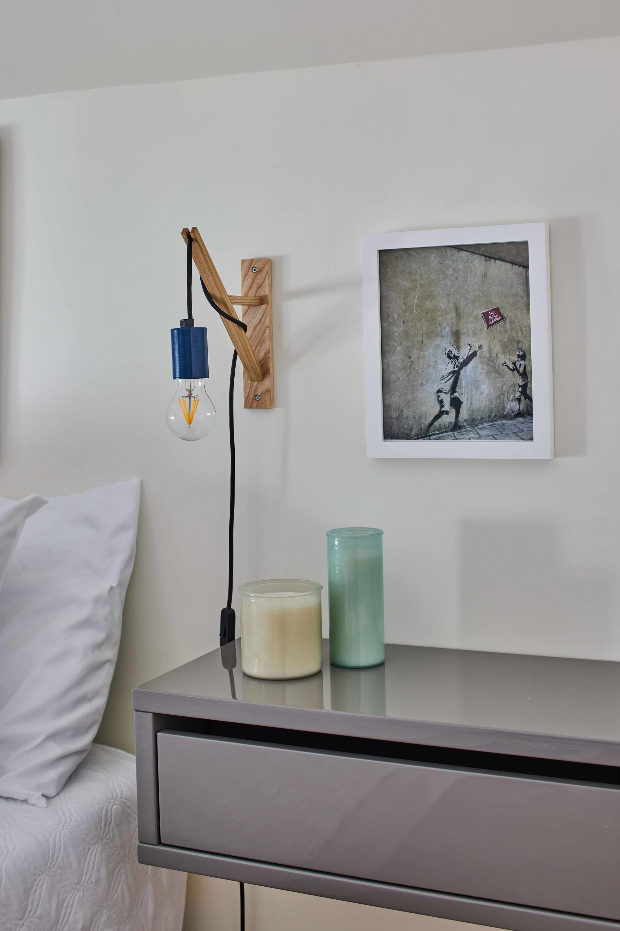 Un dormitorio con una cama, una mesita de noche con lámpara, una obra de arte enmarco y una vela verde.