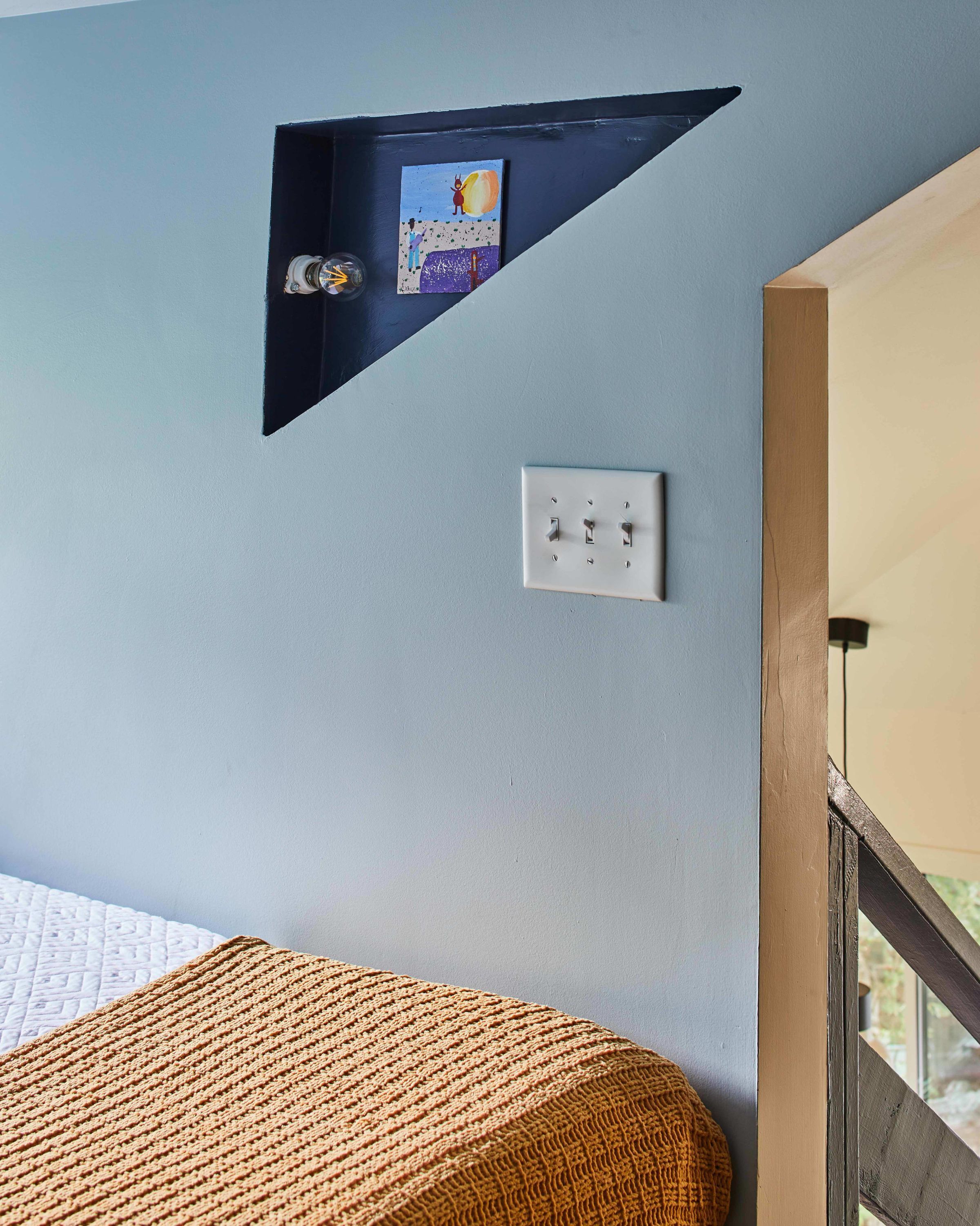 Una habitación con pared azul, un panel de interruptores blanco, un estante con un cuadro y un banco tejido.