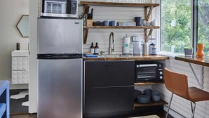 A compact kitchen with a stainless steel refrigerator, a microwave, and a sink. There is a wooden shelf above the sink, and a window providing a view of greenery outside. - Apt3 Charles - Modern Fun Sun-filled Loft (Birmingham)