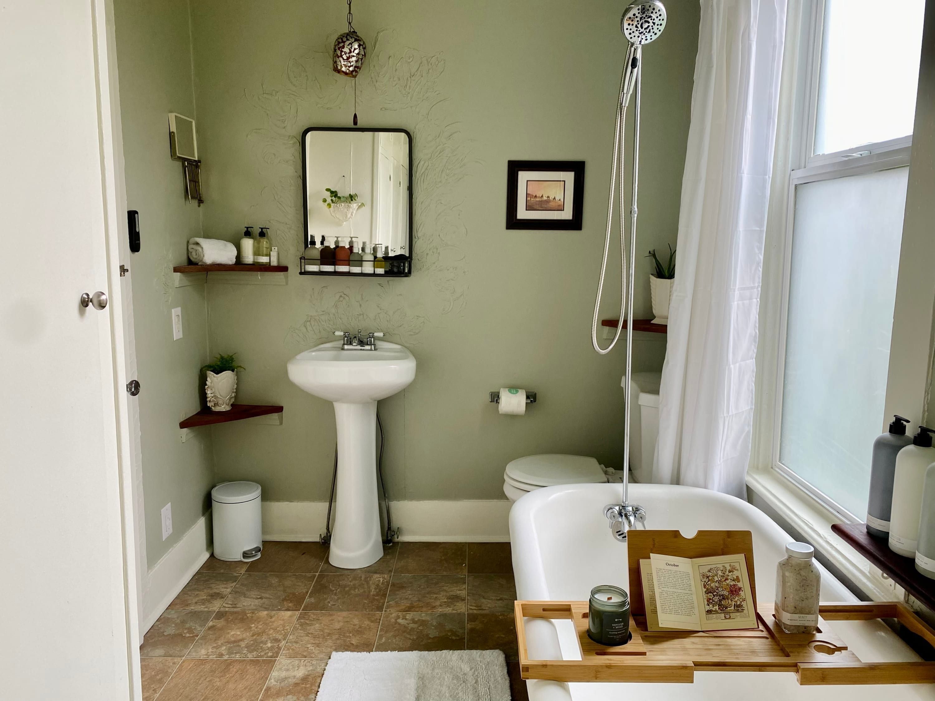 A bathroom with a white sink, toilet, and bathtub. There are shelves with toiletries and a mirror above the sink. A showerhead is mounted on the wall, and a window provides natural light.