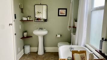 A bathroom with a white sink, toilet, and bathtub. There are shelves with toiletries and a mirror above the sink. A showerhead is mounted on the wall, and a window provides natural light.