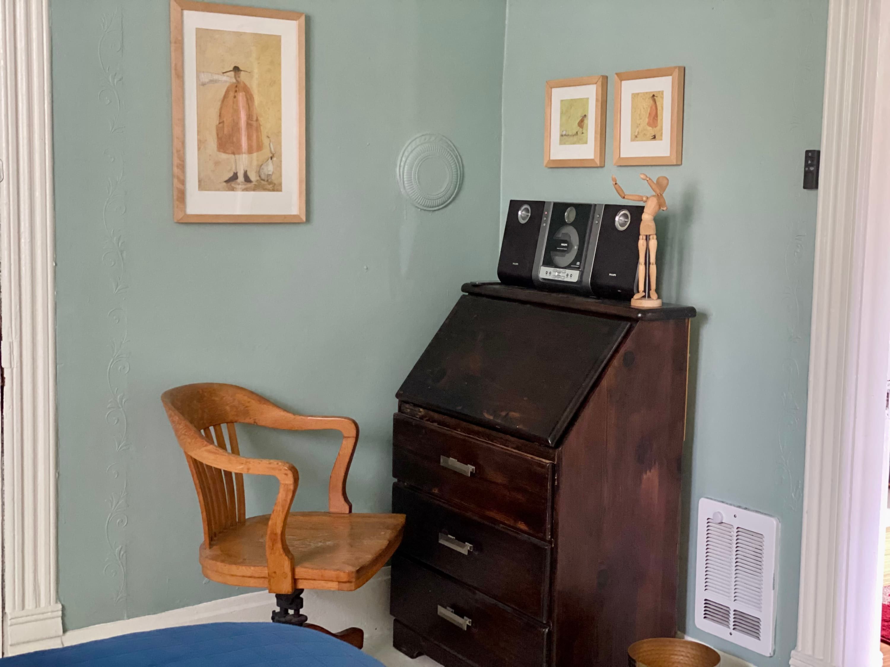 Una stanza con una sedia in legno, una scrivania scura con cassetti, opere d'arte incorniciate al muro e un tavolino con un oggetto decorativo.