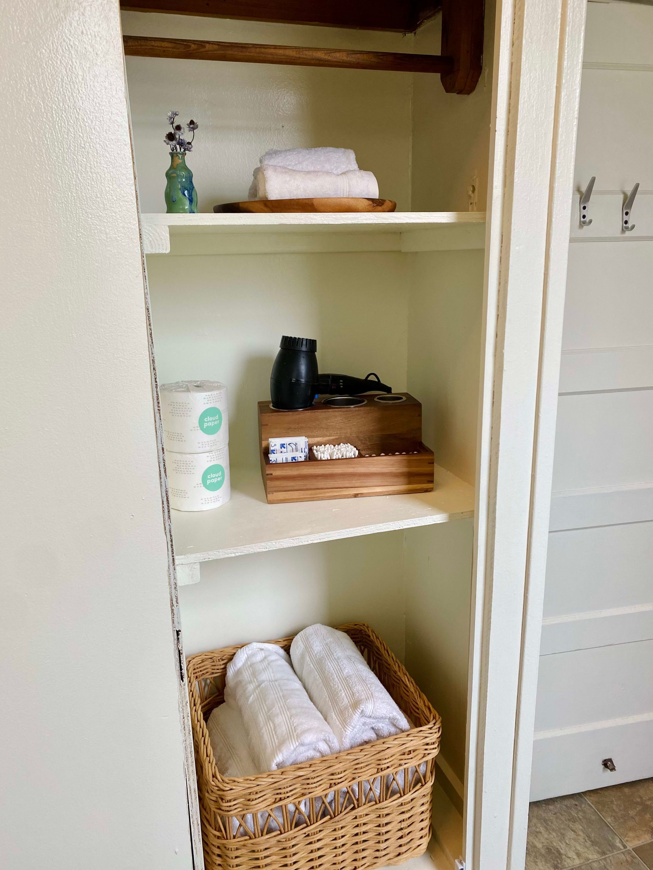 Una habitación con una pared blanca, una estantería con un florero, una cesta y un cofre de madera.