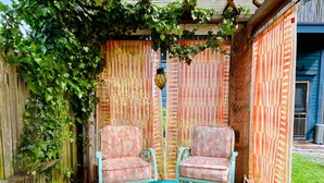 Une terrasse avec deux chaises turquoise et une table en verre, entourée d’une clôture en bois et recouverte d’un tissu à rayures orange et blanches.