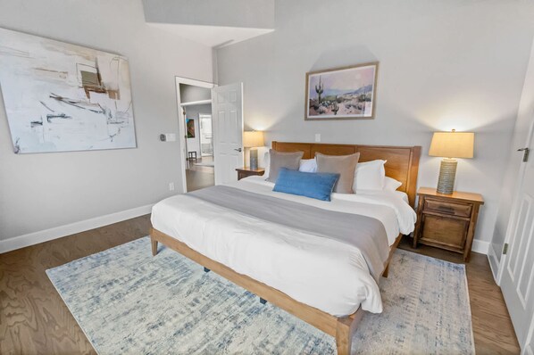 Un dormitorio con cama de madera, ropa de cama blanca, un cojín azul decorativo, una mesita de noche con lámpara, un cuadro en la pared y una obra de arte abstracta de gran tamaño.