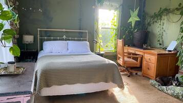 A bedroom with a bed, desk, and chair, a window with plants, and a dog bed.