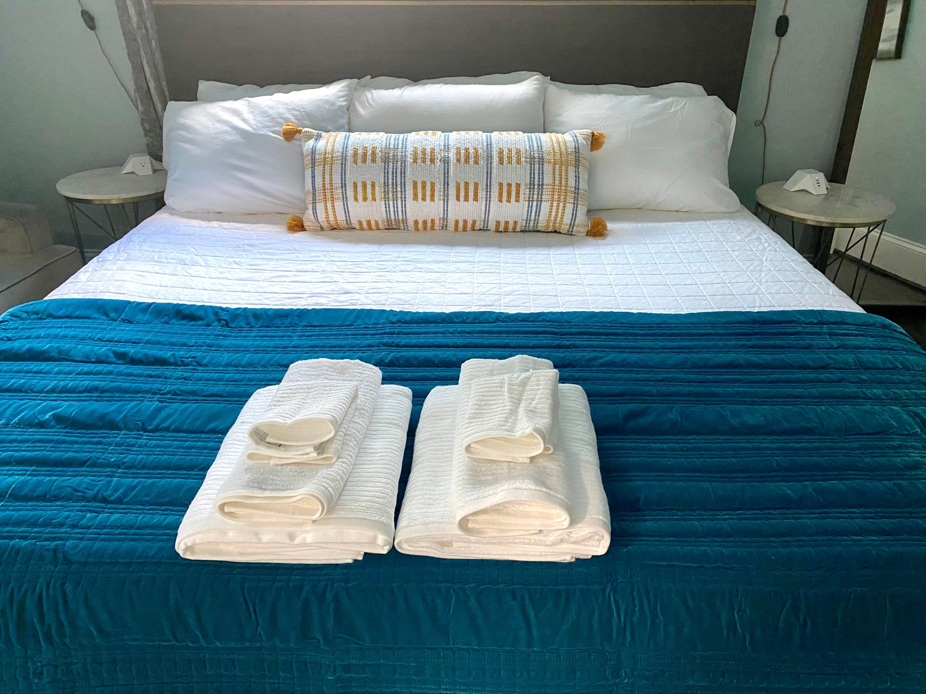 A bed with white and blue bedding, two folded towels, and a patterned pillow.